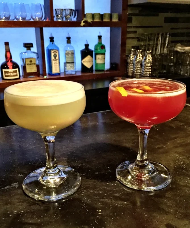 Pale yellow and pink cocktails in coupe glasses on a bar.