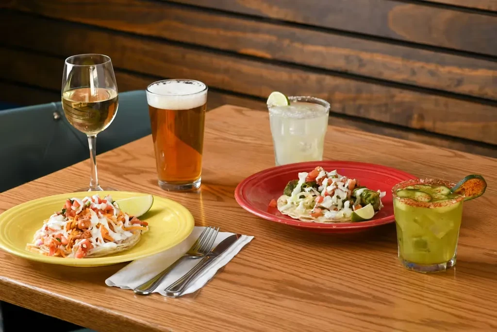 Two colorful tostadas and four drinks on a wooden restaurant table.