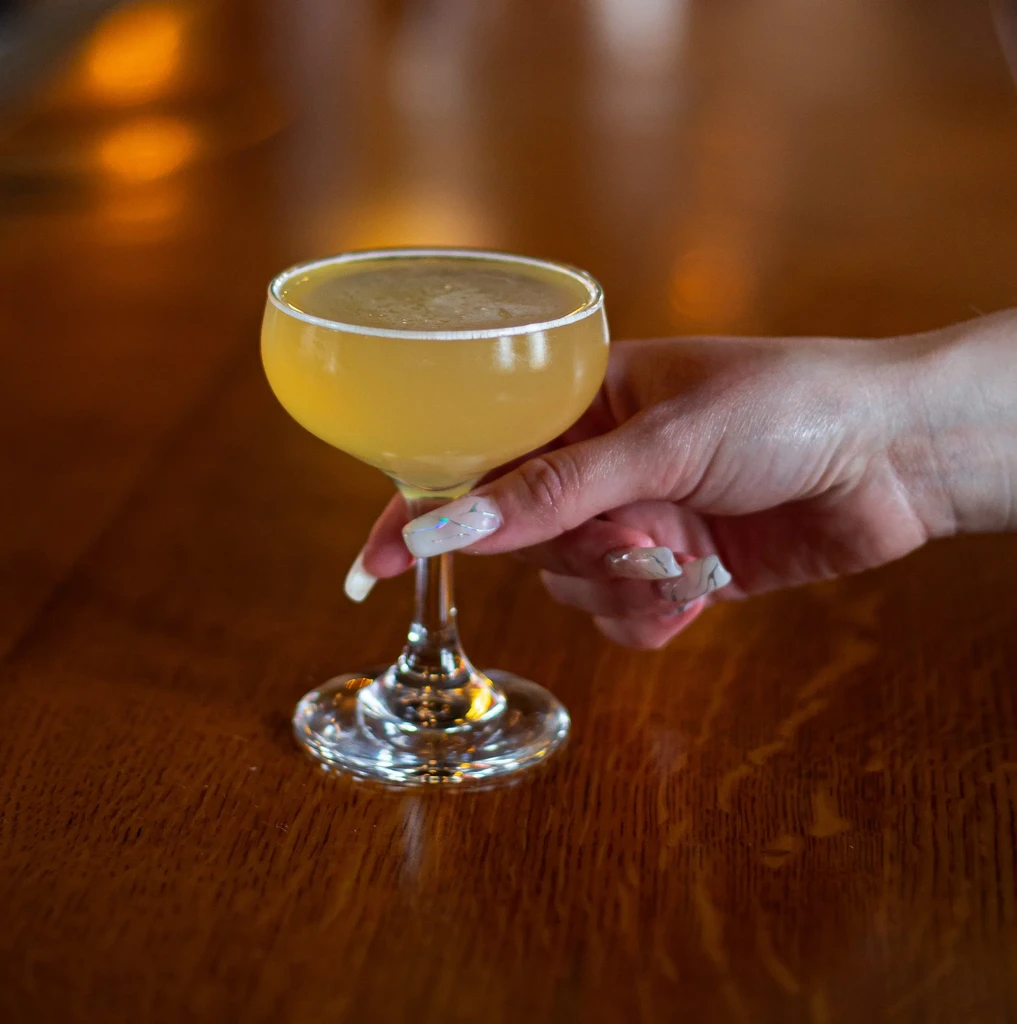 Golden-yellow cocktail in coupe glass held by hand on wooden surface.