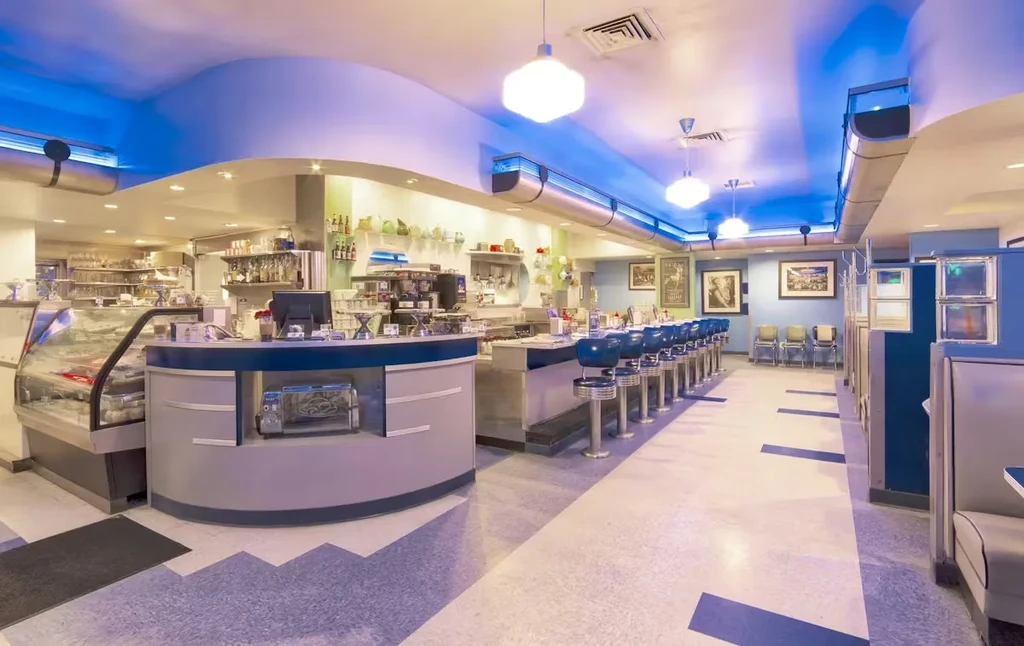 Retro diner interior with blue stools, counter, booths, and accent lighting.