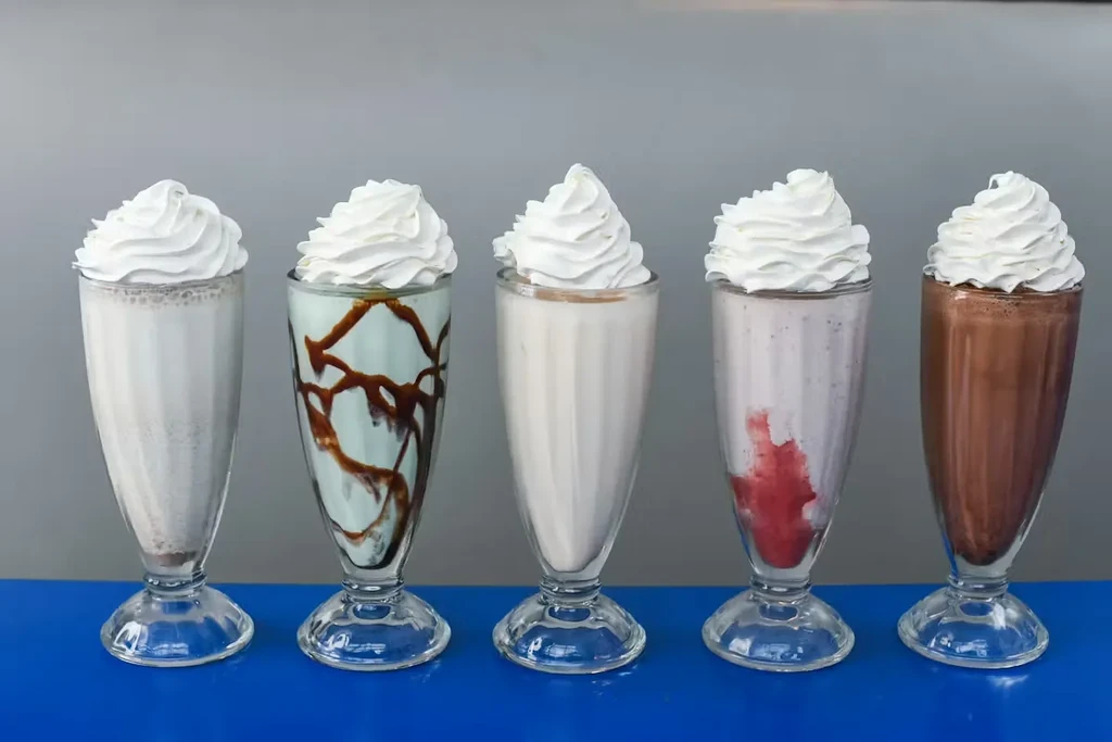 Five assorted milkshakes in clear glasses with whipped cream.