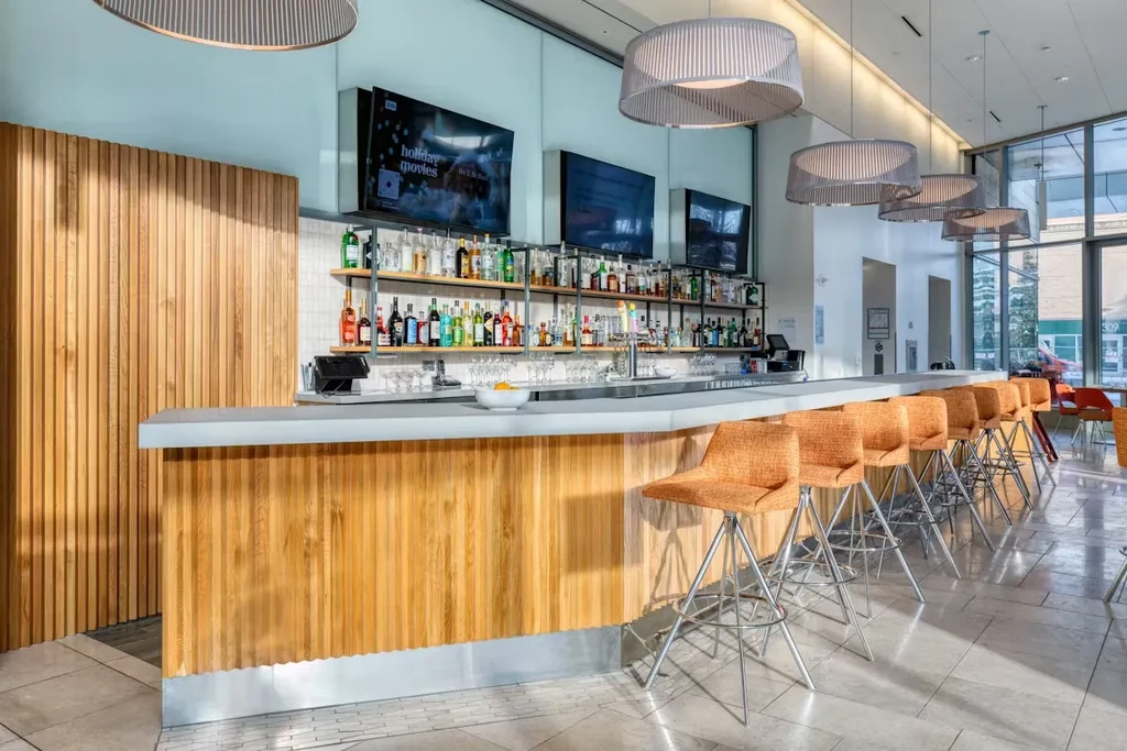 Bright modern bar with slatted counter, liquor shelves, and orange stools.