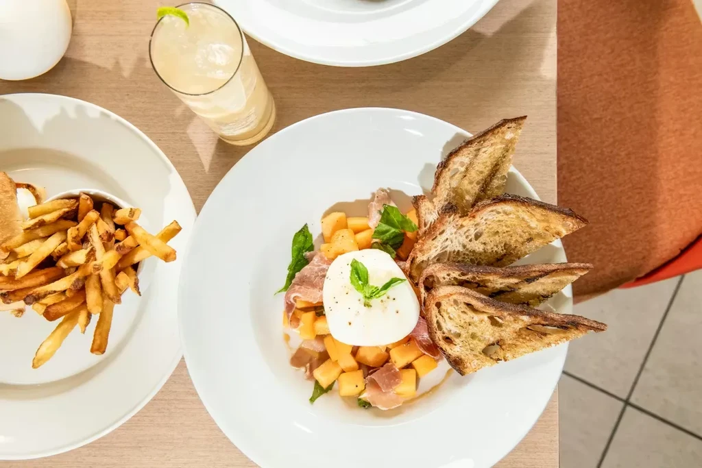 Overhead: Burrata, prosciutto, melon, bread, fries, and cocktail on table.