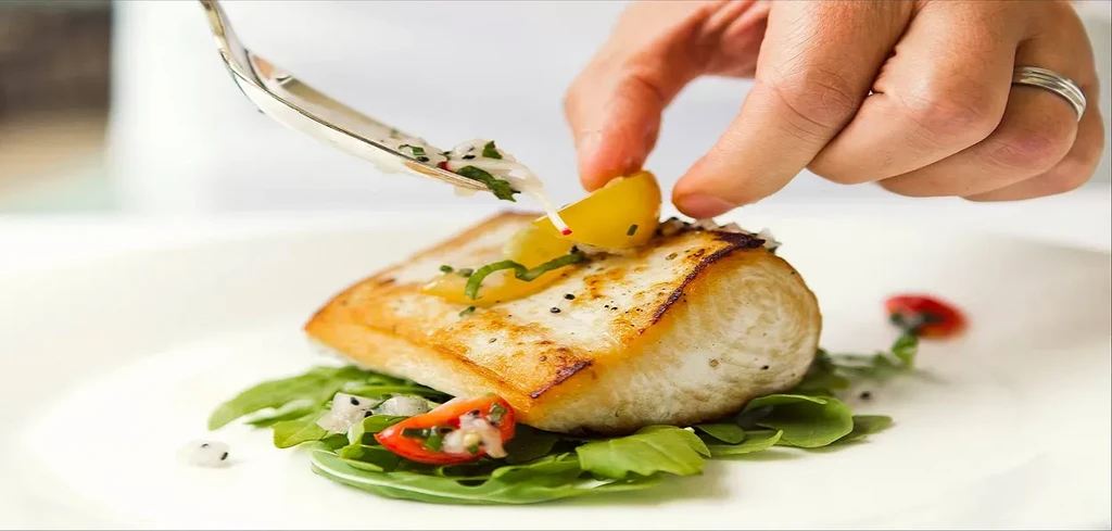 Hand plates pan-seared fish with lemon, herbs, and sauce on greens.