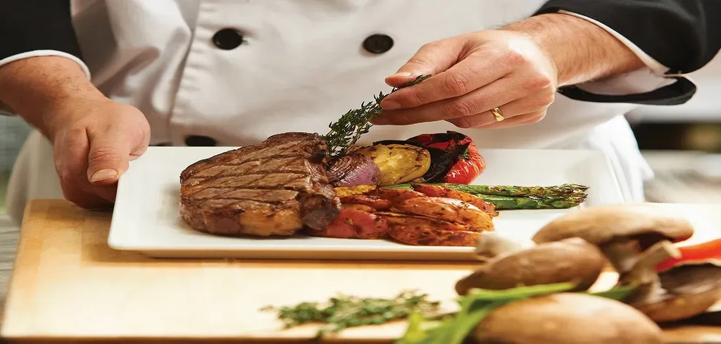 Chef plating a grilled steak with vegetables and fresh herbs.