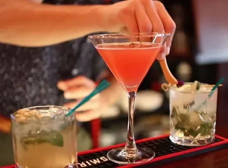 Three cocktails on a bar, a pink martini being garnished with wood.