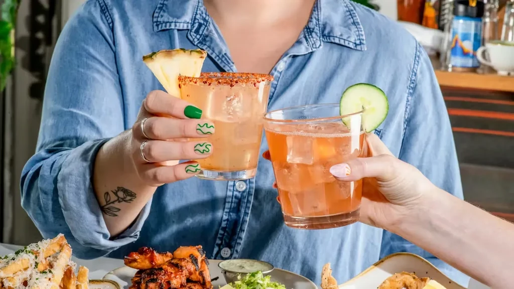 Two hands toasting drinks garnished with pineapple and cucumber.