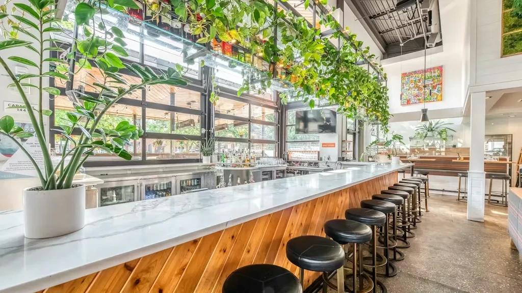 Modern bar interior with white marble counter, black stools, and greenery.