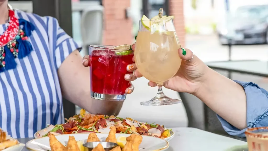 Two hands clinking cocktails, with seafood and fried food on table.