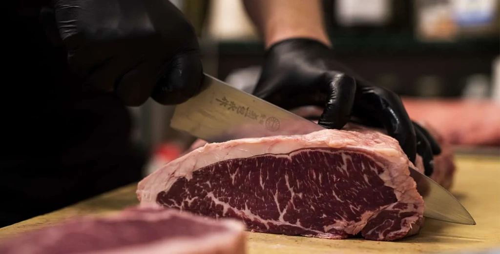 Gloved hands slicing raw, marbled beef with a Japanese chef's knife.