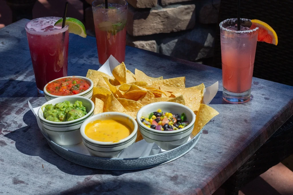 Tortilla chips, four colorful dips, and three vibrant drinks outdoors.
