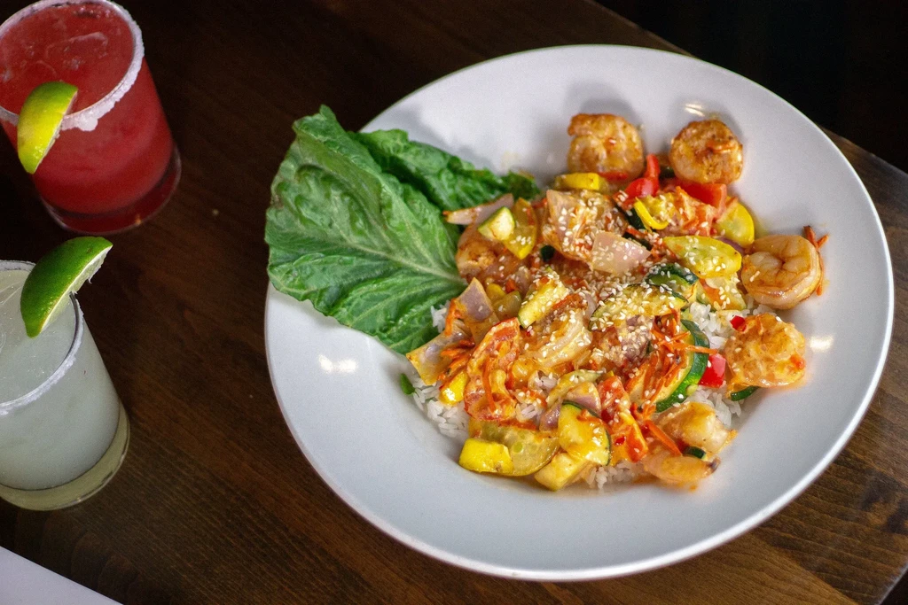 Shrimp and veggie stir-fry, two margaritas, on a wooden table.