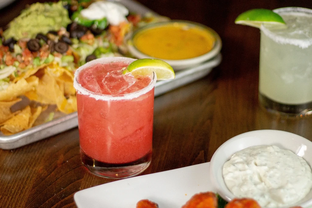 Red margarita, nachos, and various dips on a wooden table.