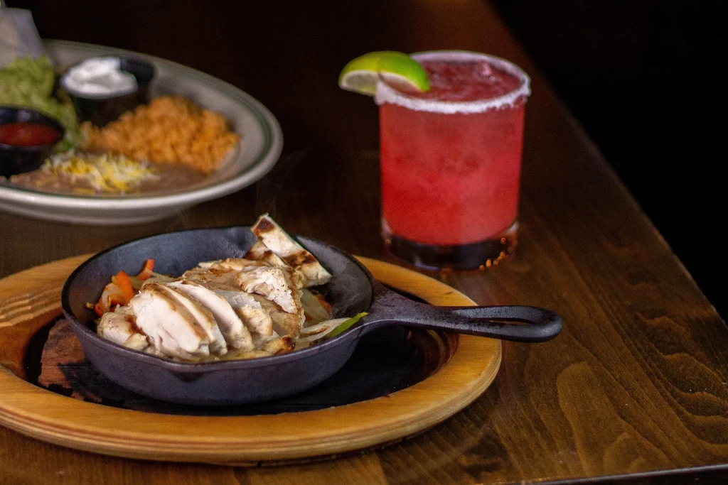 Grilled chicken fajitas skillet, sides, and a red margarita on dark wood.