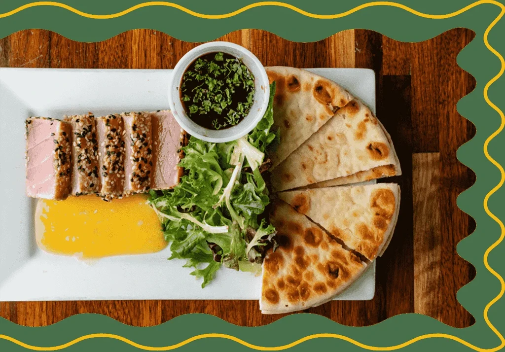Sesame-crusted seared tuna with salad, sauces, and flatbread on a wooden table.