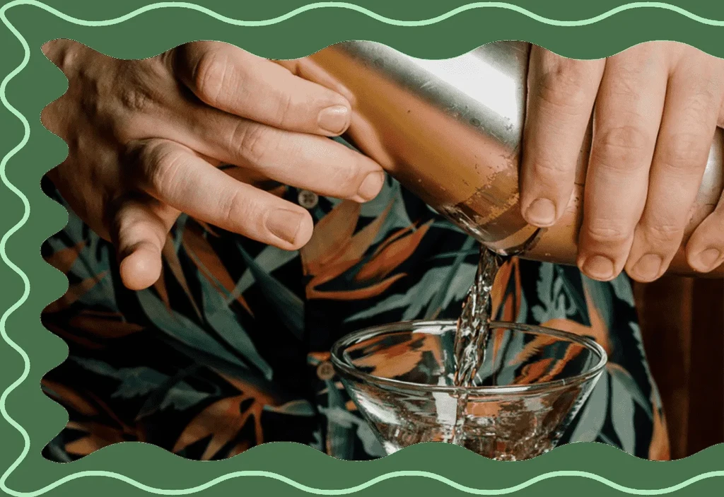 Hands pour cocktail from shaker into martini glass, tropical shirt visible.
