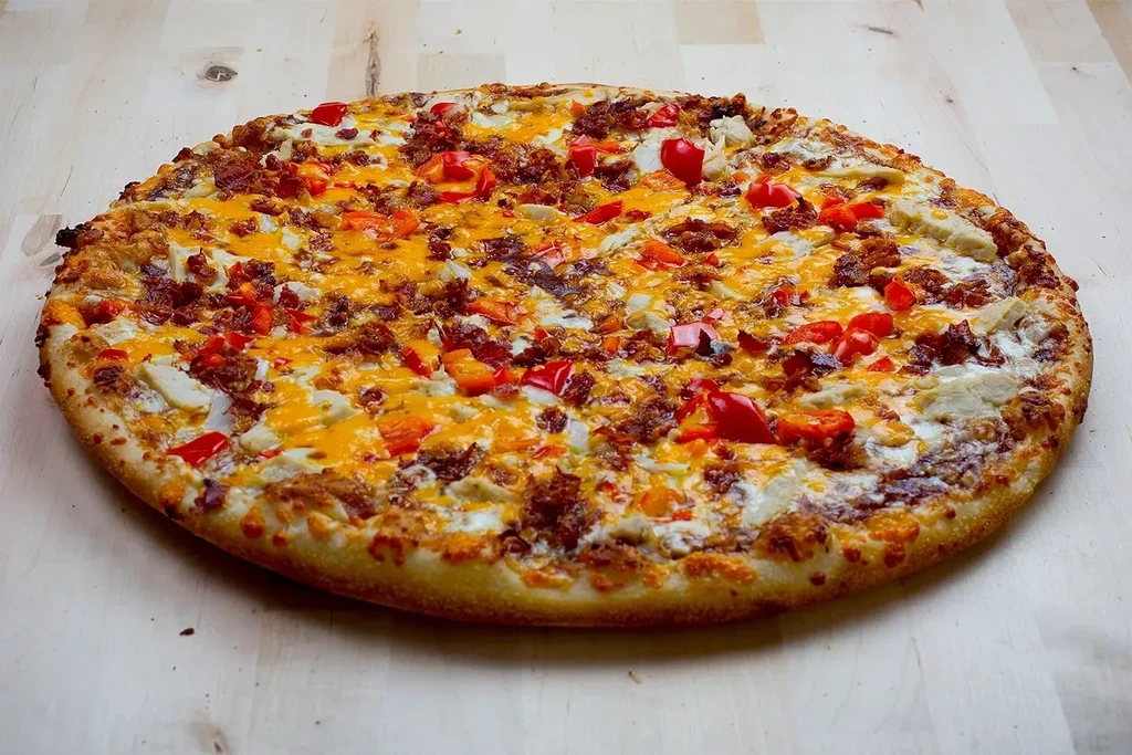 Round pizza with meats, cheeses, red and orange peppers on wood.