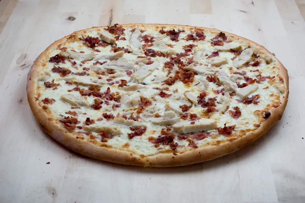 Oval pizza with creamy white sauce, chicken, and bacon on wood.