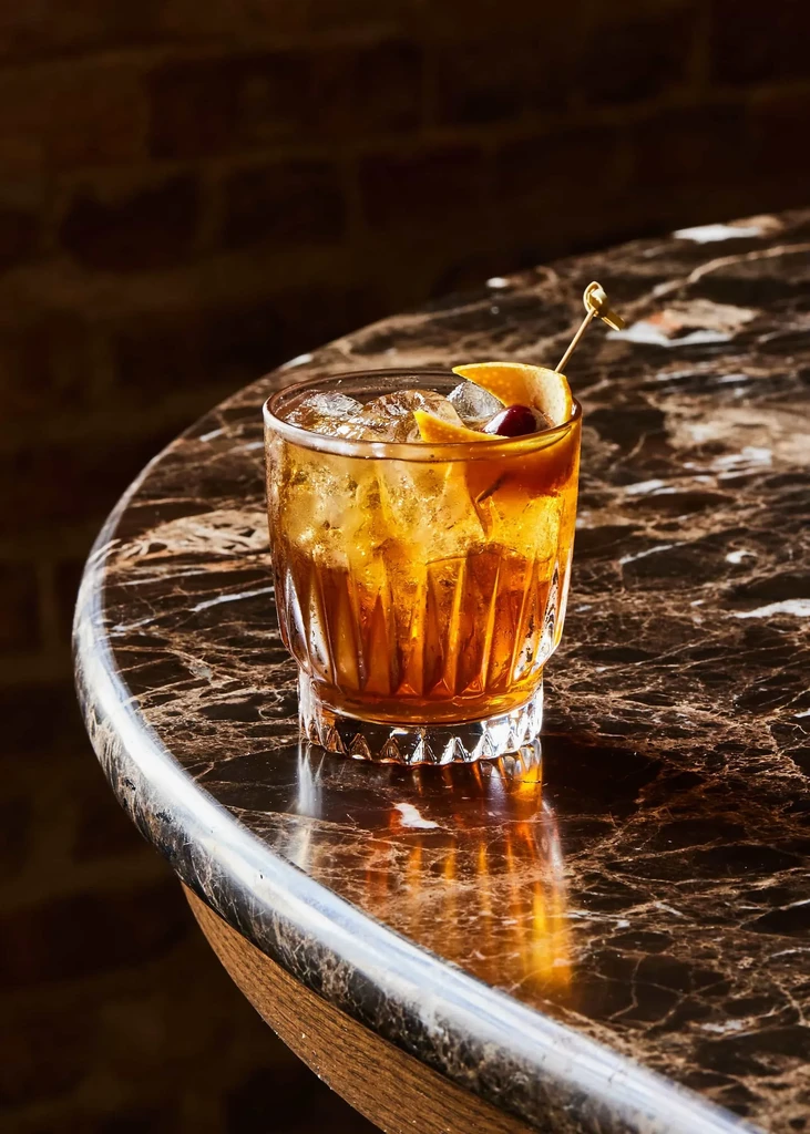 Amber cocktail with orange peel and cherry in faceted glass on marble.