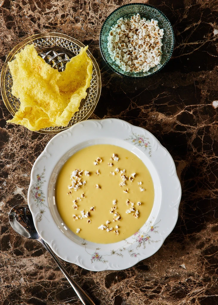 Top-down: Yellow soup garnished with popcorn, crackers, and a spoon.