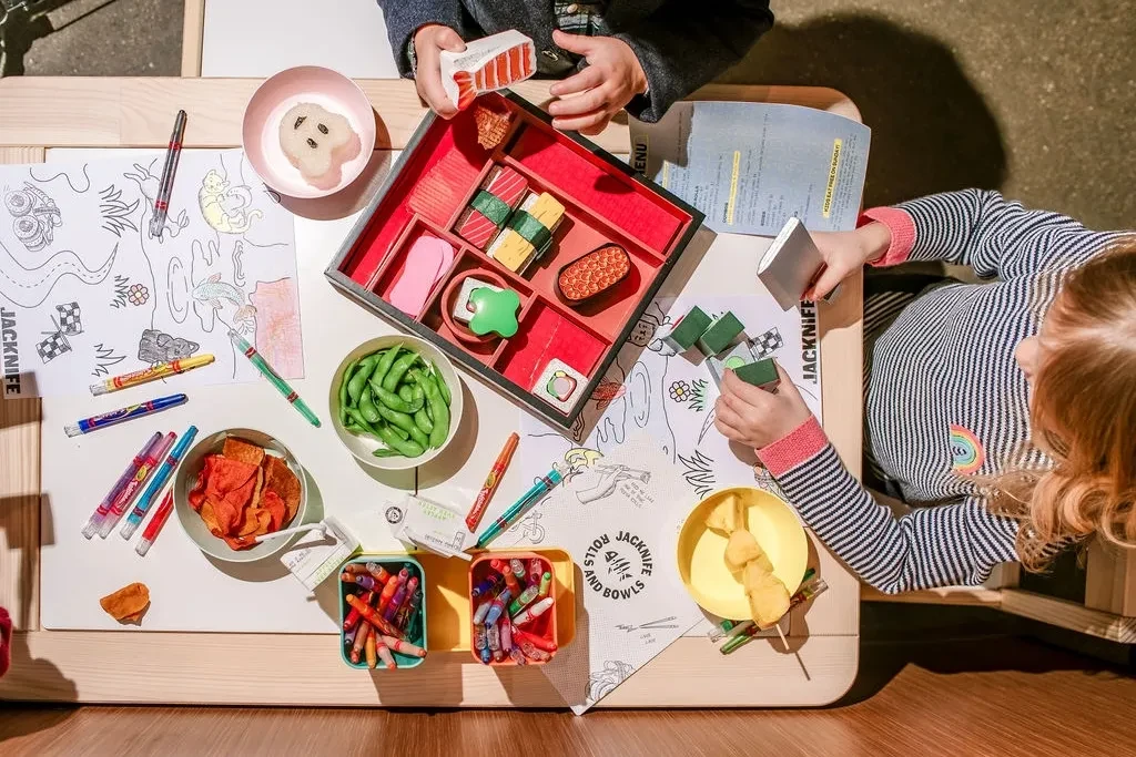 Children's hands play with toy sushi, blocks, and food at a table.