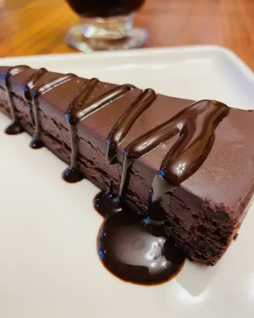 Dark rectangular chocolate cake with glossy ganache and sauce on plate.