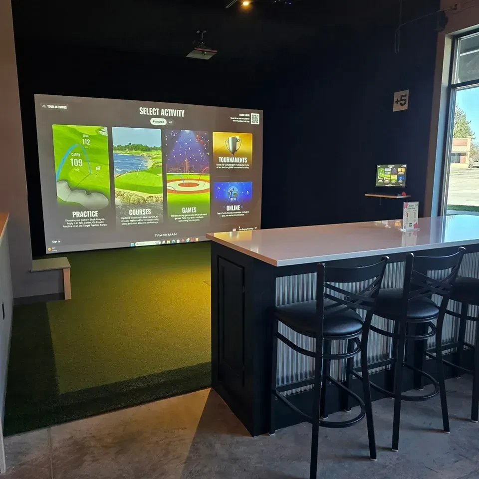 Indoor golf simulator bay with large screen, bar, and hitting mat.