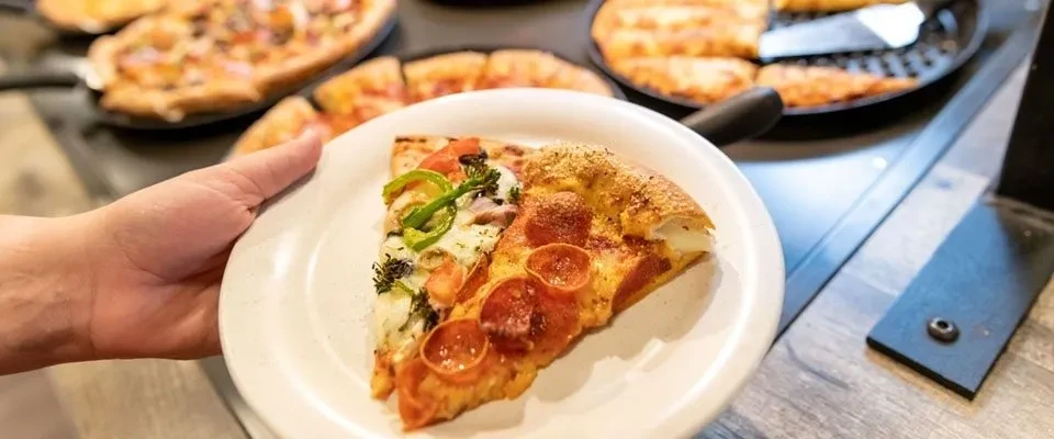 Two pizza slices, pepperoni and vegetable, held over blurred buffet pizzas.
