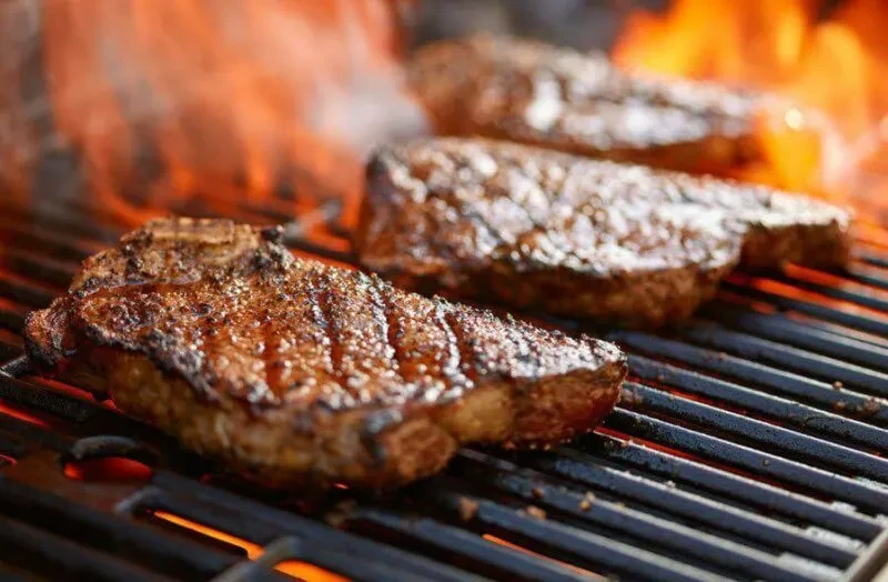 Three thick steaks grilling on a metal grate over bright flames.