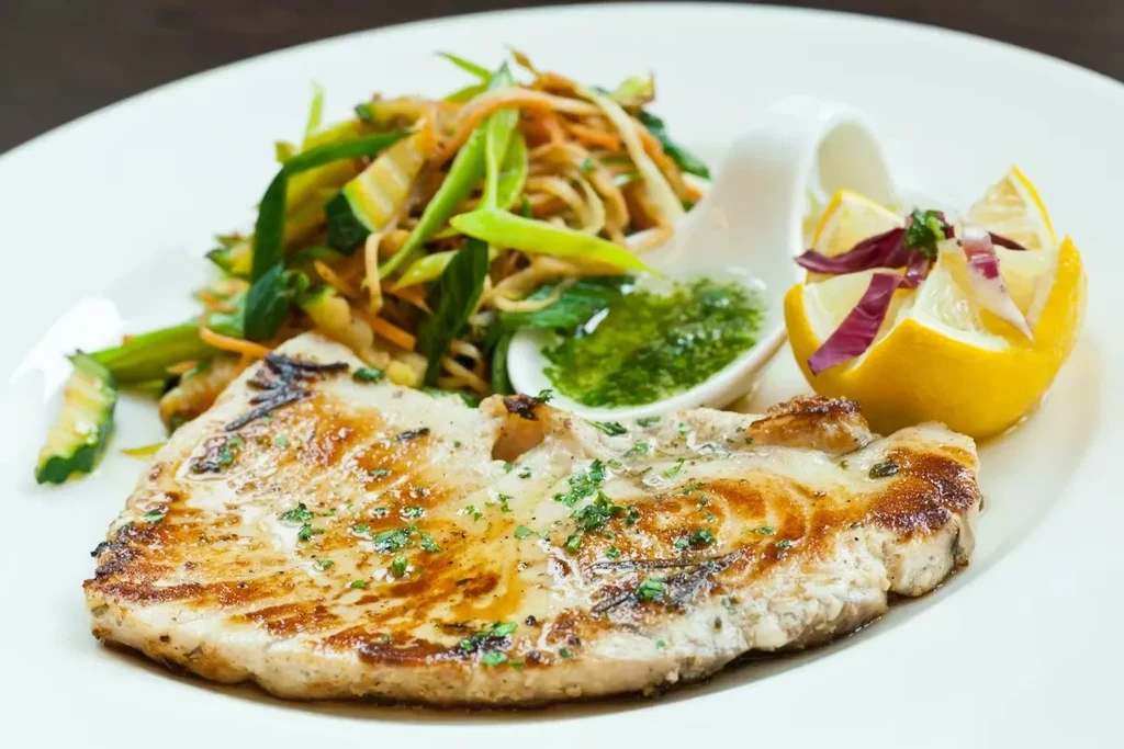 Professionally plated grilled meat with julienned vegetables, green sauce, and lemon.