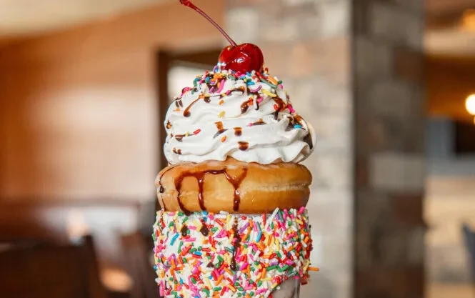 Elaborate milkshake with sprinkles, donut, whipped cream, chocolate, and a cherry.