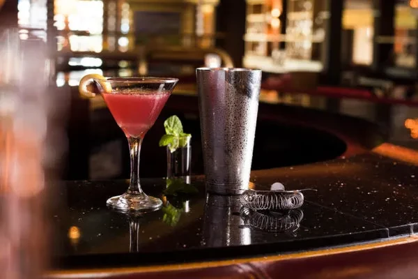 Pink martini cocktail and bar tools on a dark reflective bar.