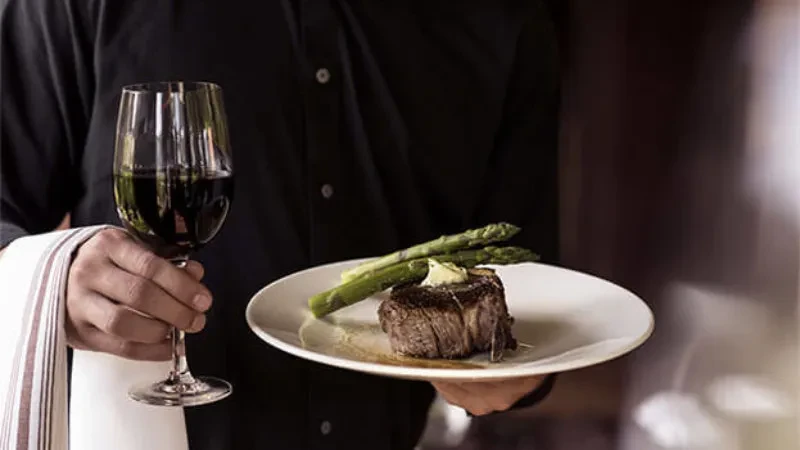 Close-up: Server presents steak, asparagus, and red wine.