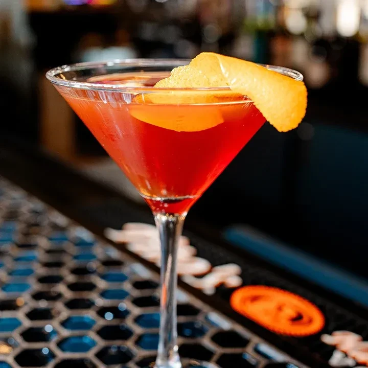 Red-orange cocktail in martini glass with citrus twist on tiled counter.