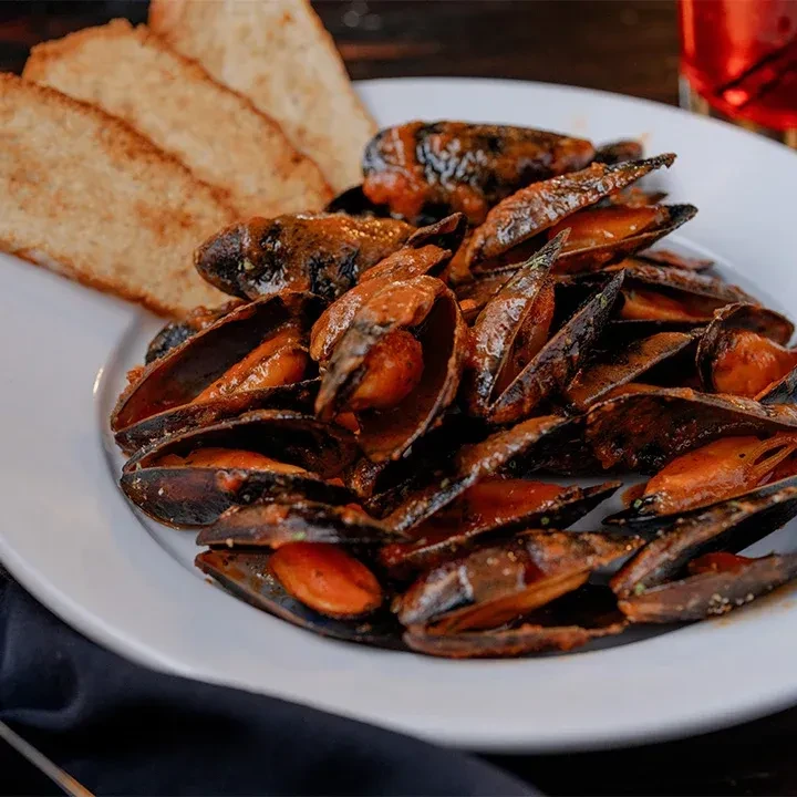 Mussels in reddish-brown sauce with toasted bread on a white plate.