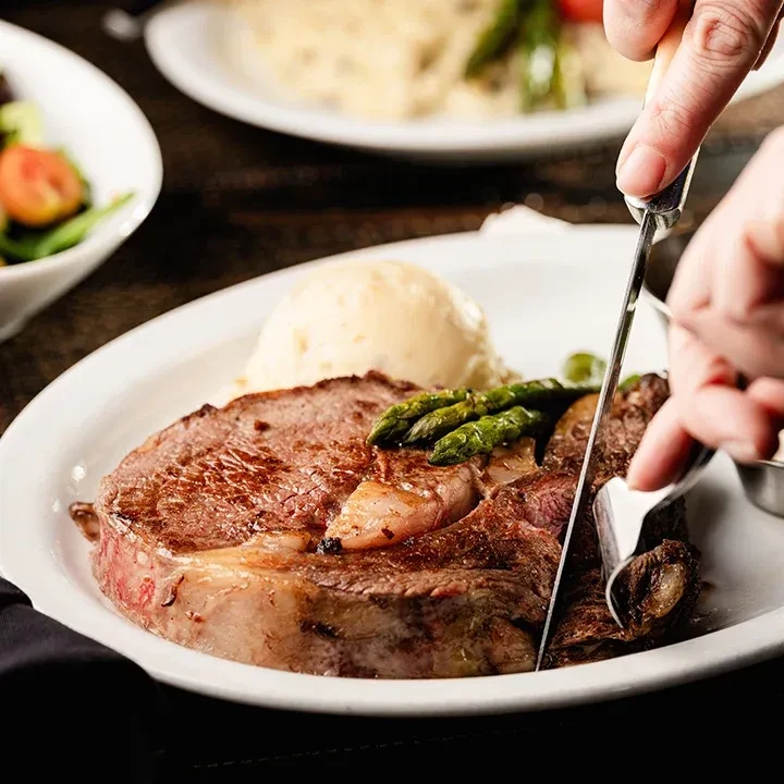 Hands cutting a seared medium-rare steak, mashed potatoes, and asparagus.