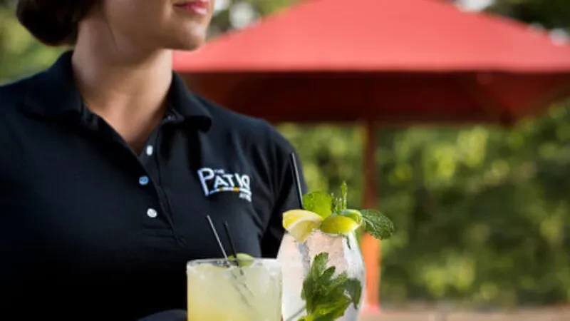 Server holds a mojito and light-colored drink on a patio.
