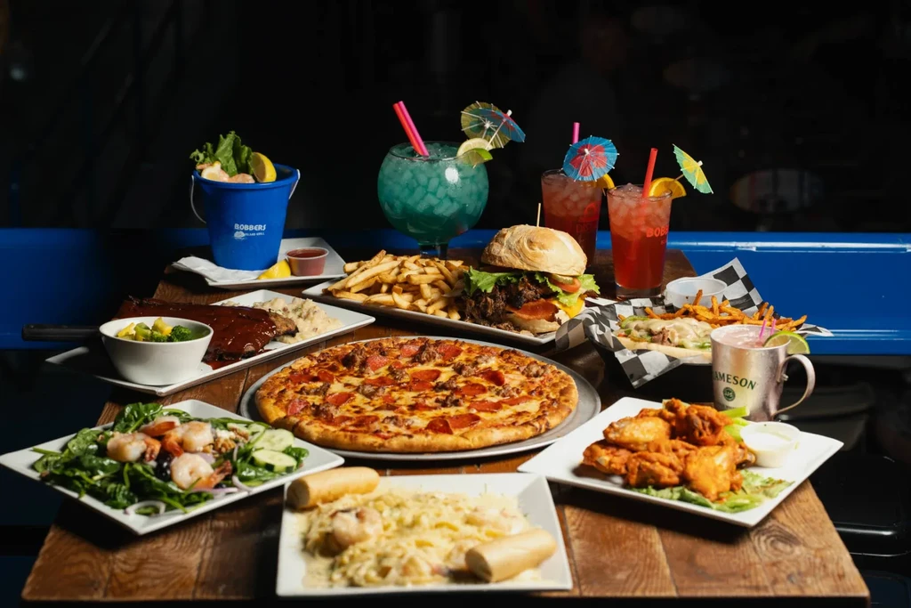 Rustic table laden with pizza, ribs, burgers, pasta, wings, and cocktails.