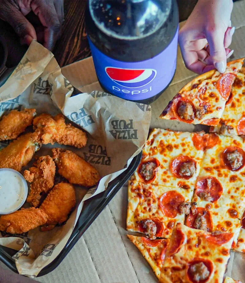 Hand lifts pepperoni pizza slice, fried chicken wings, and Pepsi on table.