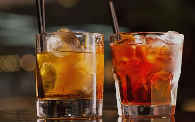 Two chilled amber and reddish-orange cocktails with ice, straws, citrus.