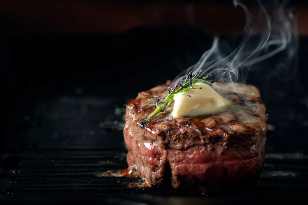 Hot, thick grilled steak with melting butter, rosemary on grate.