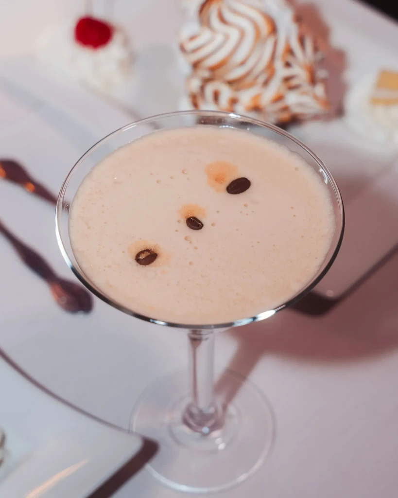 Frothy, creamy cocktail with coffee beans in martini glass, blurred desserts.
