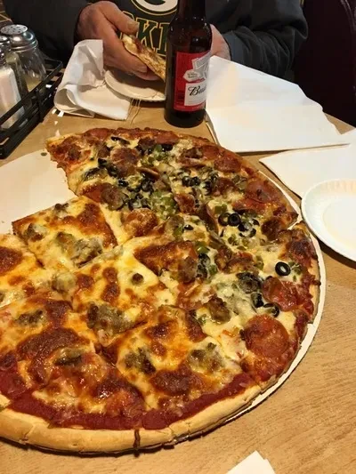 Overhead half-and-half pizza, slice held, with Budweiser bottle on wooden table.