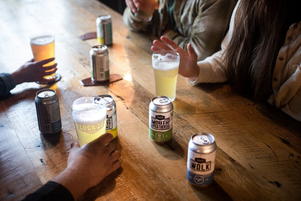 Craft beer cans and glasses on a wooden table with people's hands.