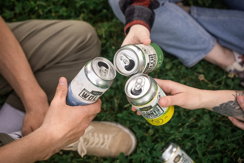 Top-down view: three hands holding open branded beer cans outdoors.