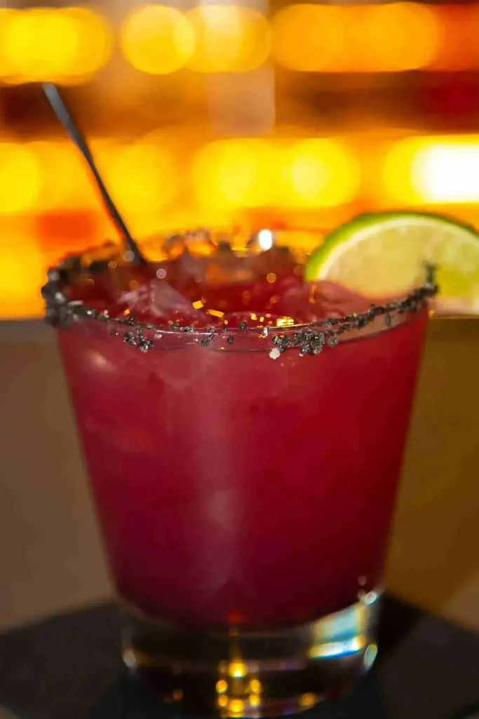 Red cocktail in glass with dark rim, lime, ice, and bokeh.