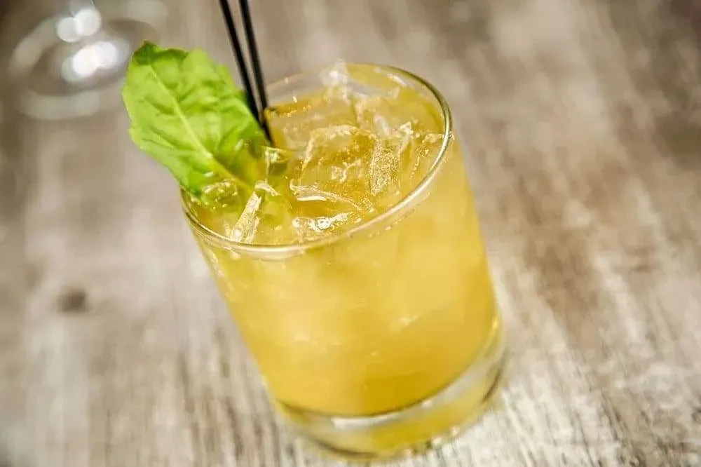 Yellowish-green cocktail in tumbler with ice, green leaf, black straws.