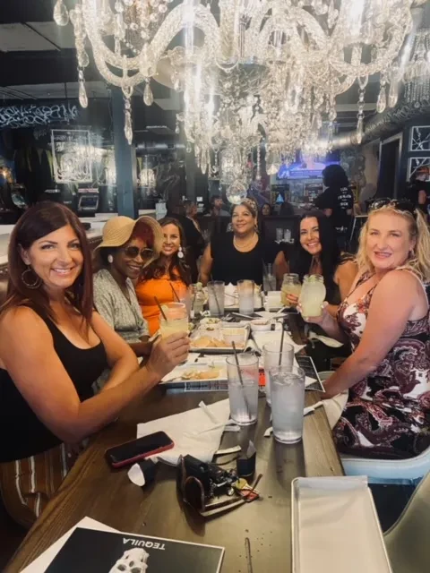 Six smiling women pose with drinks at a restaurant table with chandeliers.