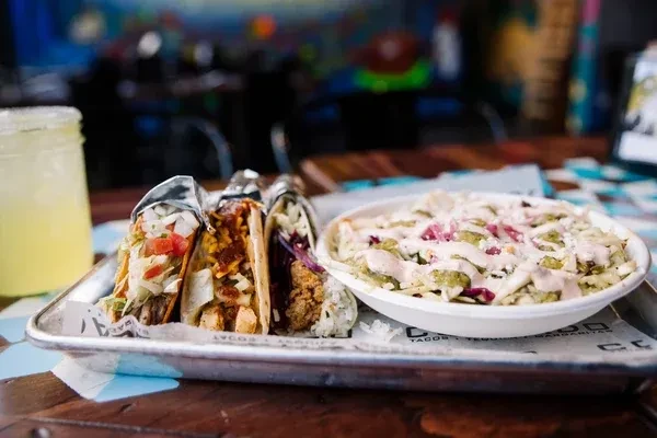 Foil-wrapped tacos, side dish, and yellow drink on a metal tray.