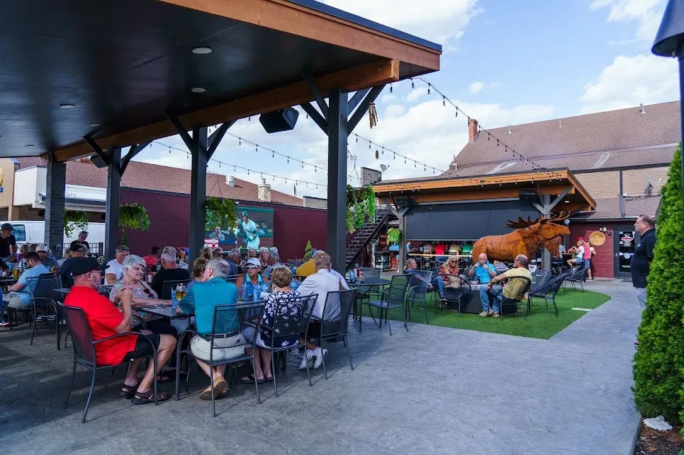 Lively outdoor patio with patrons, moose sculpture, baseball game, and string lights.